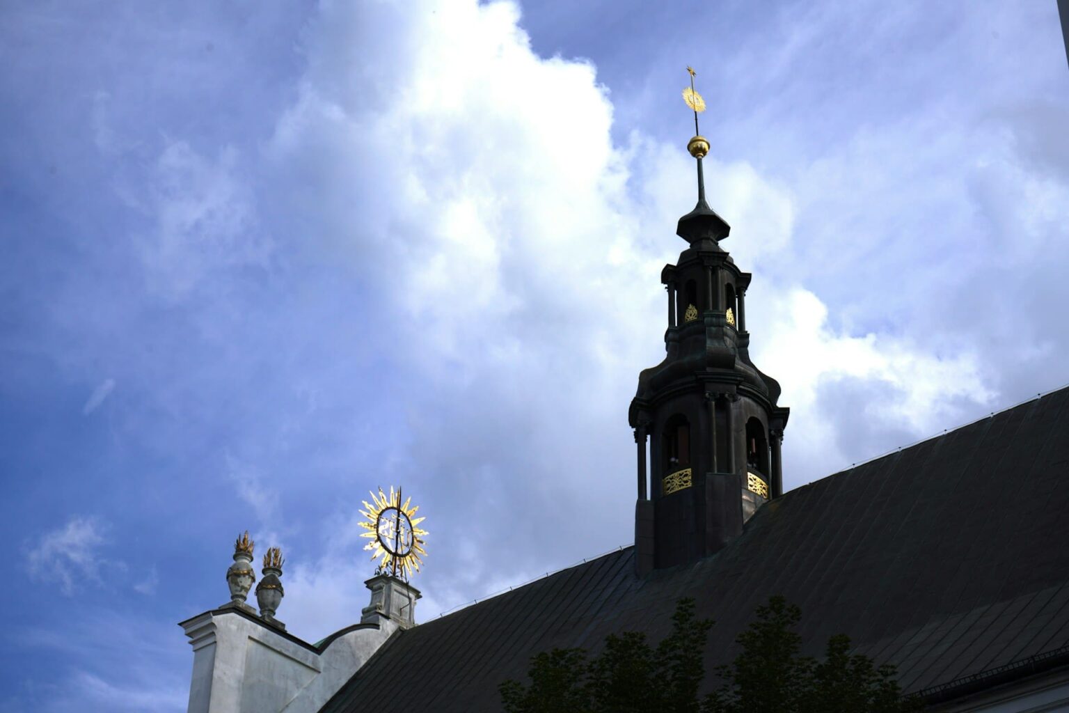 Sufixos (sufiksoj) em esperanto Torre escura com detalhes dourados e um sol dourado no alto de uma igreja, sob céu azul com nuvens