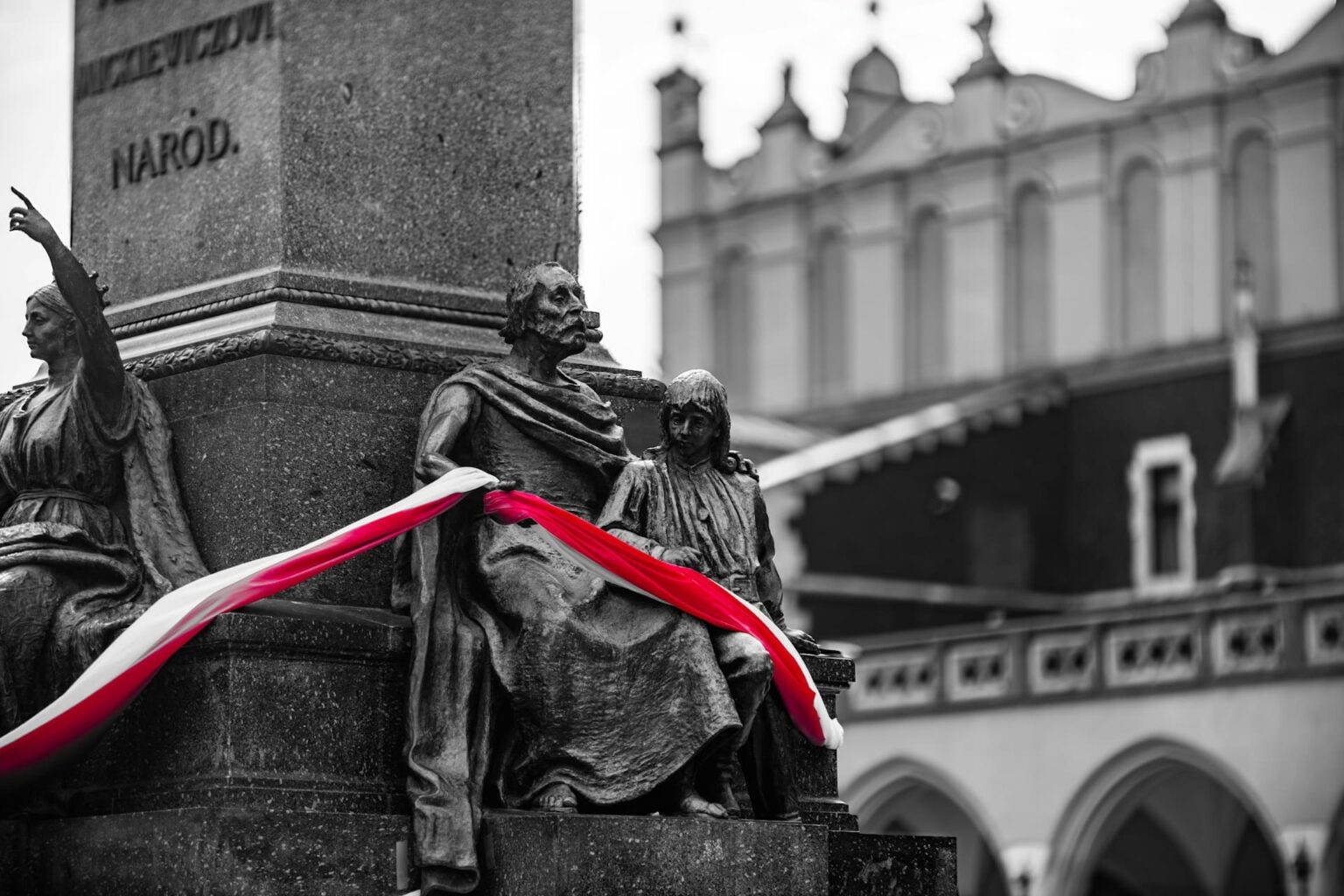 Monumento de bronze com uma faixa branca e vermelha sobre as estátuas, numa foto em preto e branco em que só a faixa tem cor