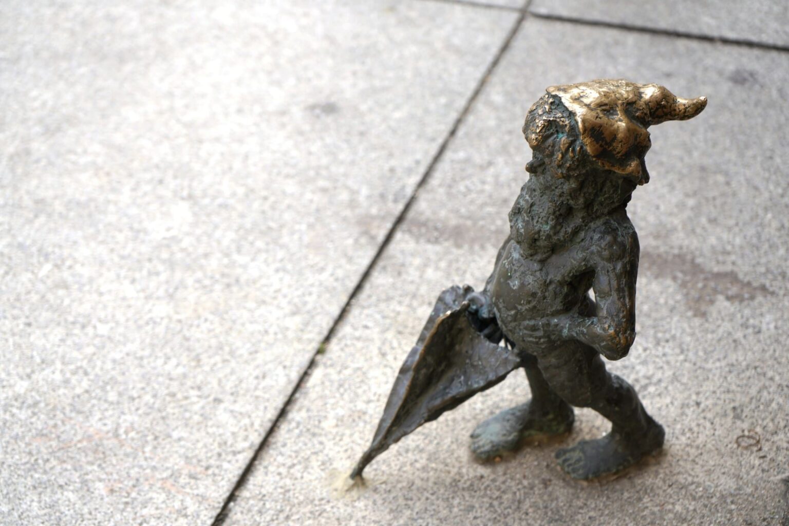 Pequena estátua de bronze de um anão barbudo de gorro, segurando um guarda-chuva fechado sobre a calçada