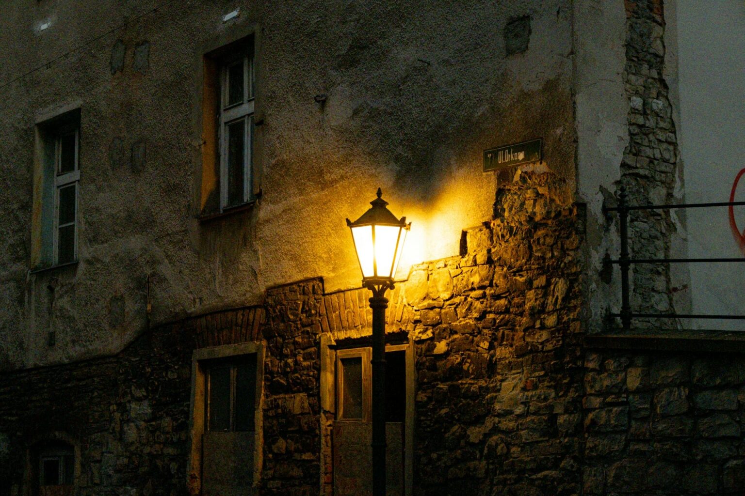Gênero (genro) em esperanto Poste de luz antigo aceso ao anoitecer diante da fachada desgastada de um prédio de pedra e reboco
