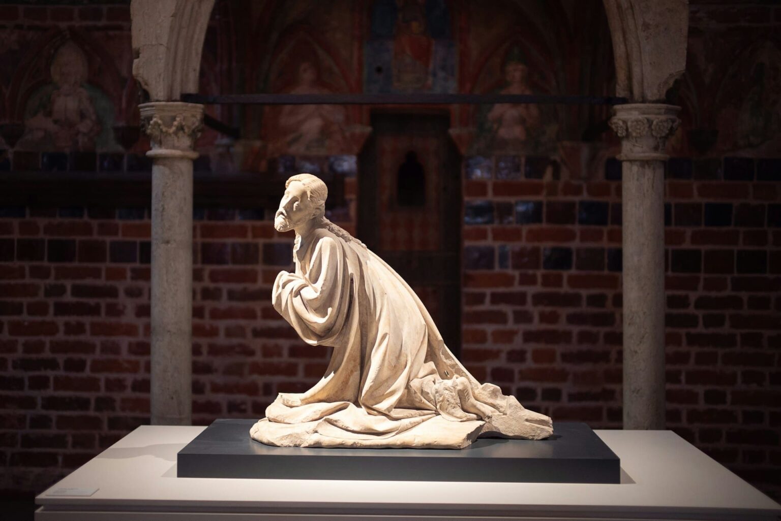 Perguntas (demandoj) em esperanto Estátua de pedra clara de um homem barbado ajoelhado, com as mãos juntas, num museu com paredes de tijolo e arcos com pinturas antigas