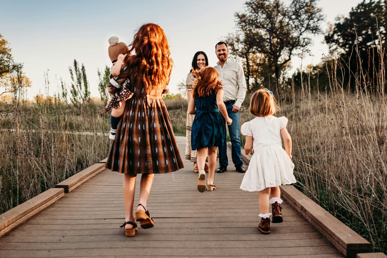 Família num caminho de tábuas no campo: duas meninas correm em direção aos pais, e uma moça leva um bebê no colo