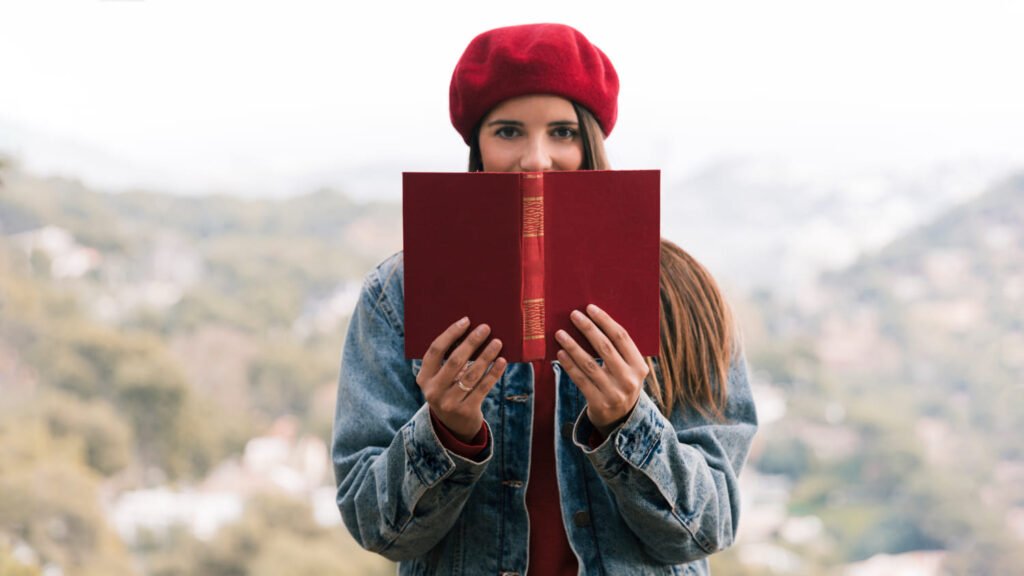 Mulher de boina vermelha e jaqueta jeans esconde metade do rosto atrás de um livro vermelho