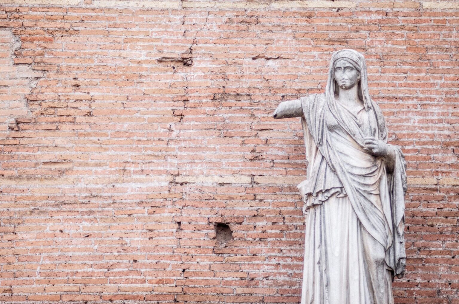 Estátua de mulher em pedra com o braço estendido, diante de um muro de tijolos