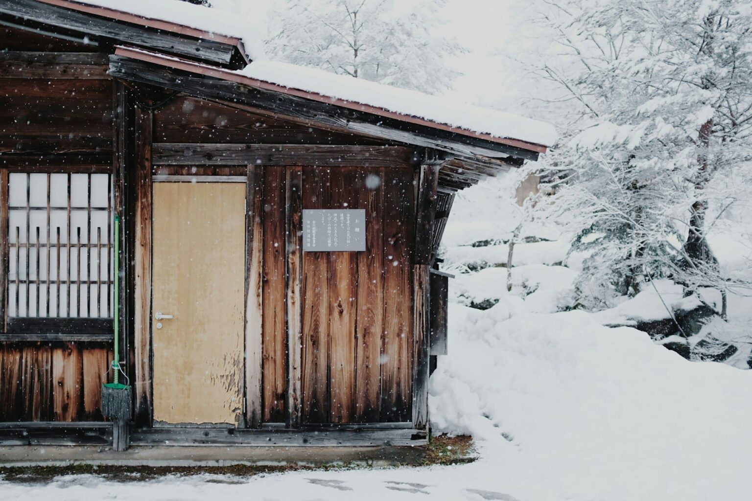 Casa antiga de madeira com porta clara e janela de treliça, com neve no telhado e neve caindo