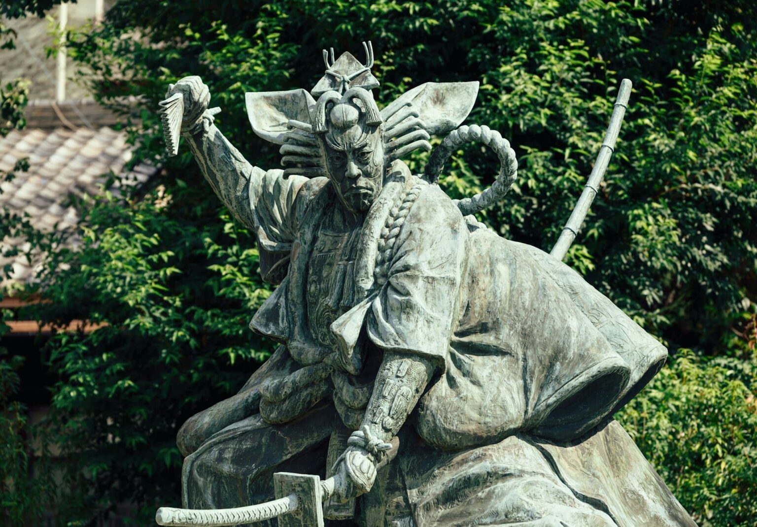 Estátua de bronze de um guerreiro em pose dramática, com espada e leque, diante de árvores