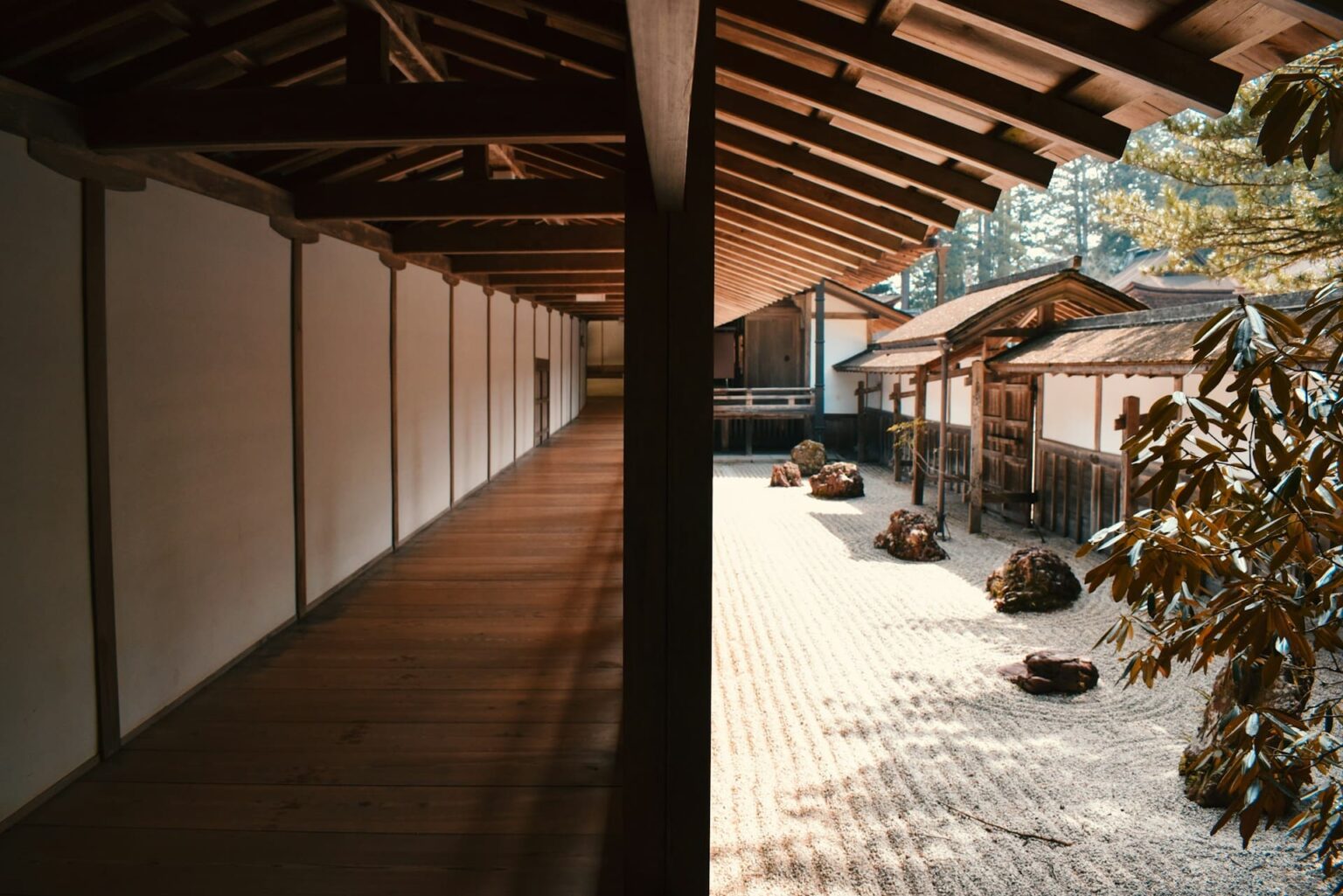 Corredor de madeira coberto ao lado de um jardim de pedras e areia rastelada, com pinheiros ao fundo