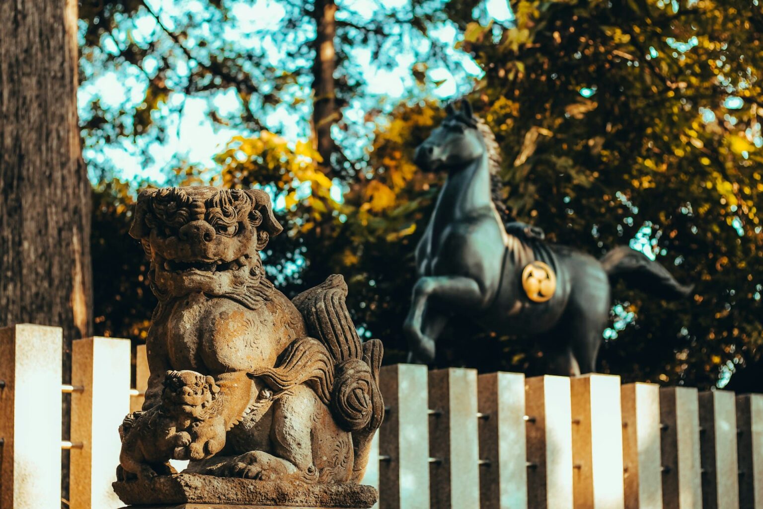Estátua de pedra de cão-leão guardião com um filhote, diante de um cavalo de bronze e árvores de outono