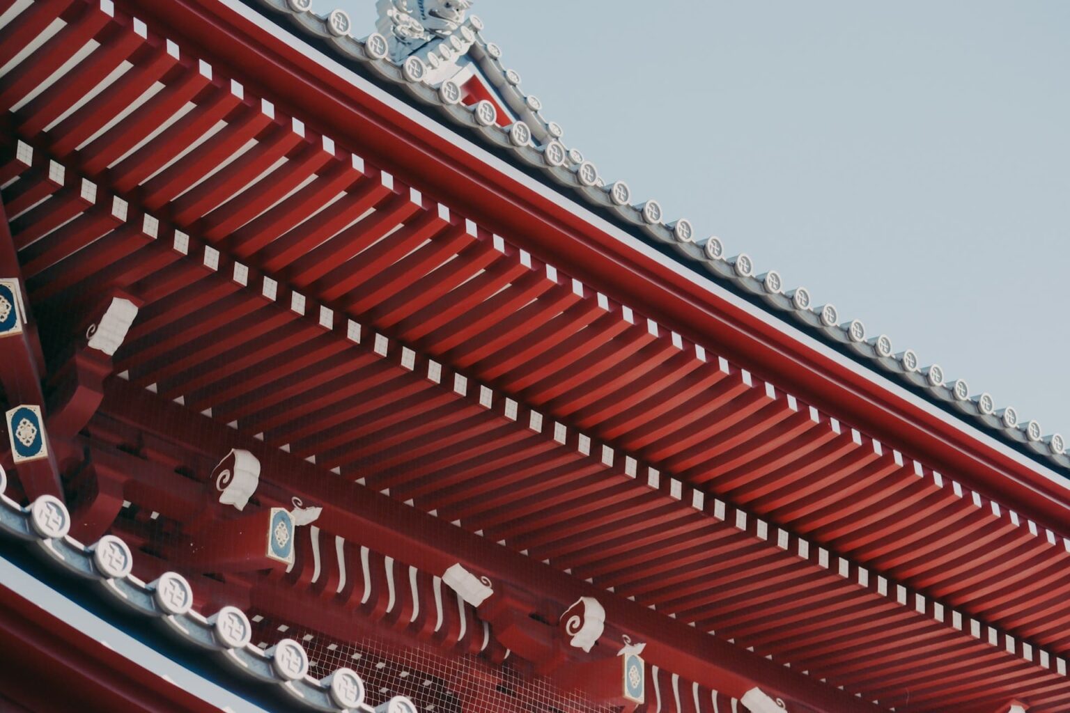 Beiral vermelho de templo com caibros pintados e telhas cinza com emblemas, contra o céu azul