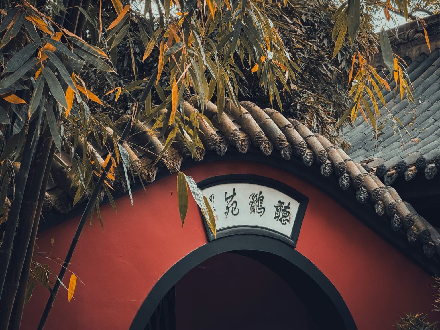 Arco redondo num muro vermelho, com placa de caligrafia, sob telhado de telhas cinza e folhas de bambu