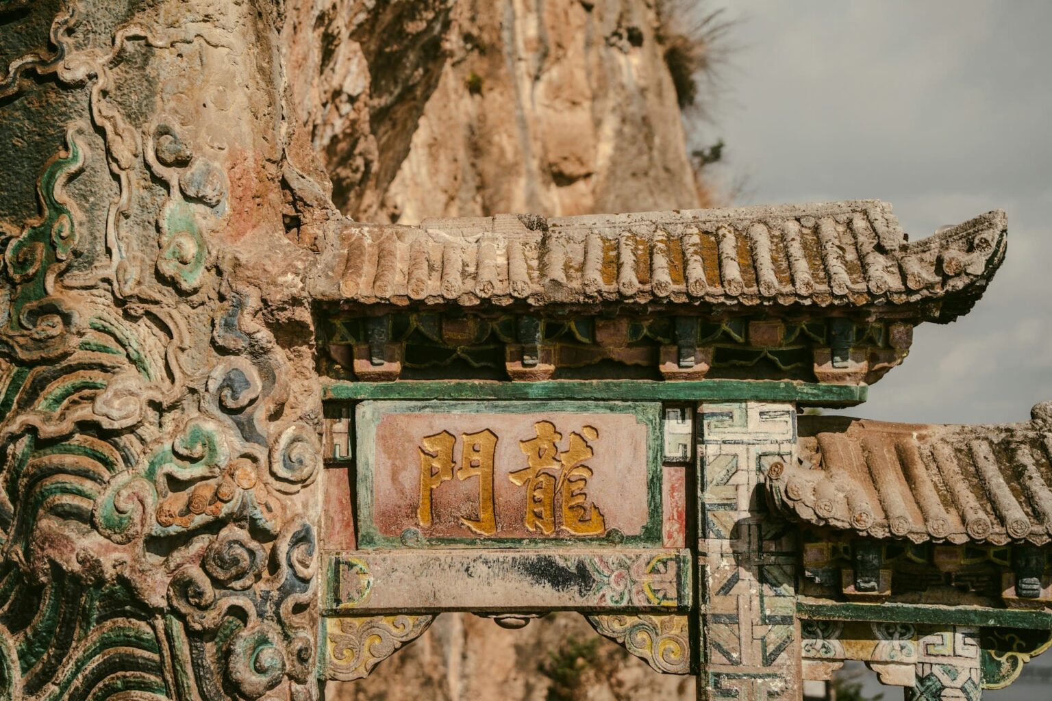 Portal de pedra com telhado de telhas e a inscrição dourada 龍門, junto a um paredão de rocha
