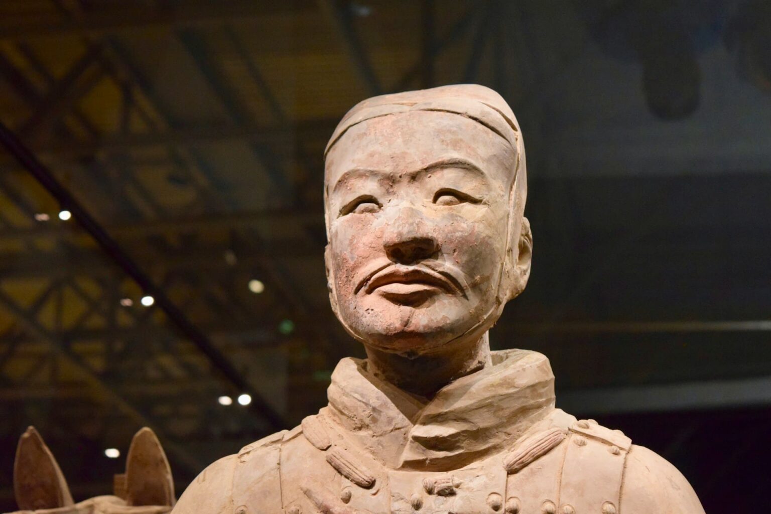 Rosto de um guerreiro de terracota, de bigode e expressão séria, sob a luz de um museu