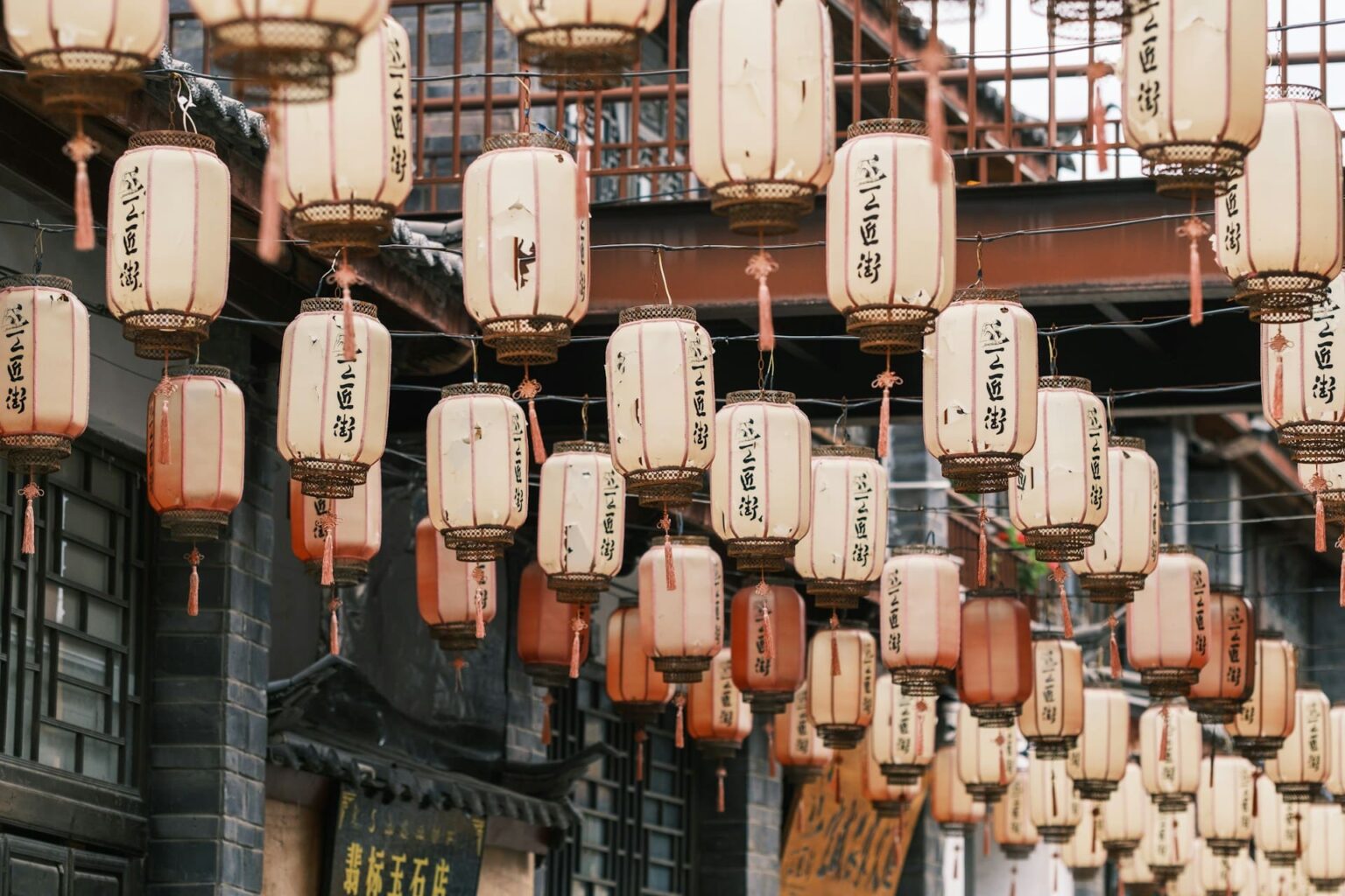 Lanternas de papel cor de creme e alaranjadas com caracteres chineses, penduradas em fileiras sobre uma rua de casas antigas