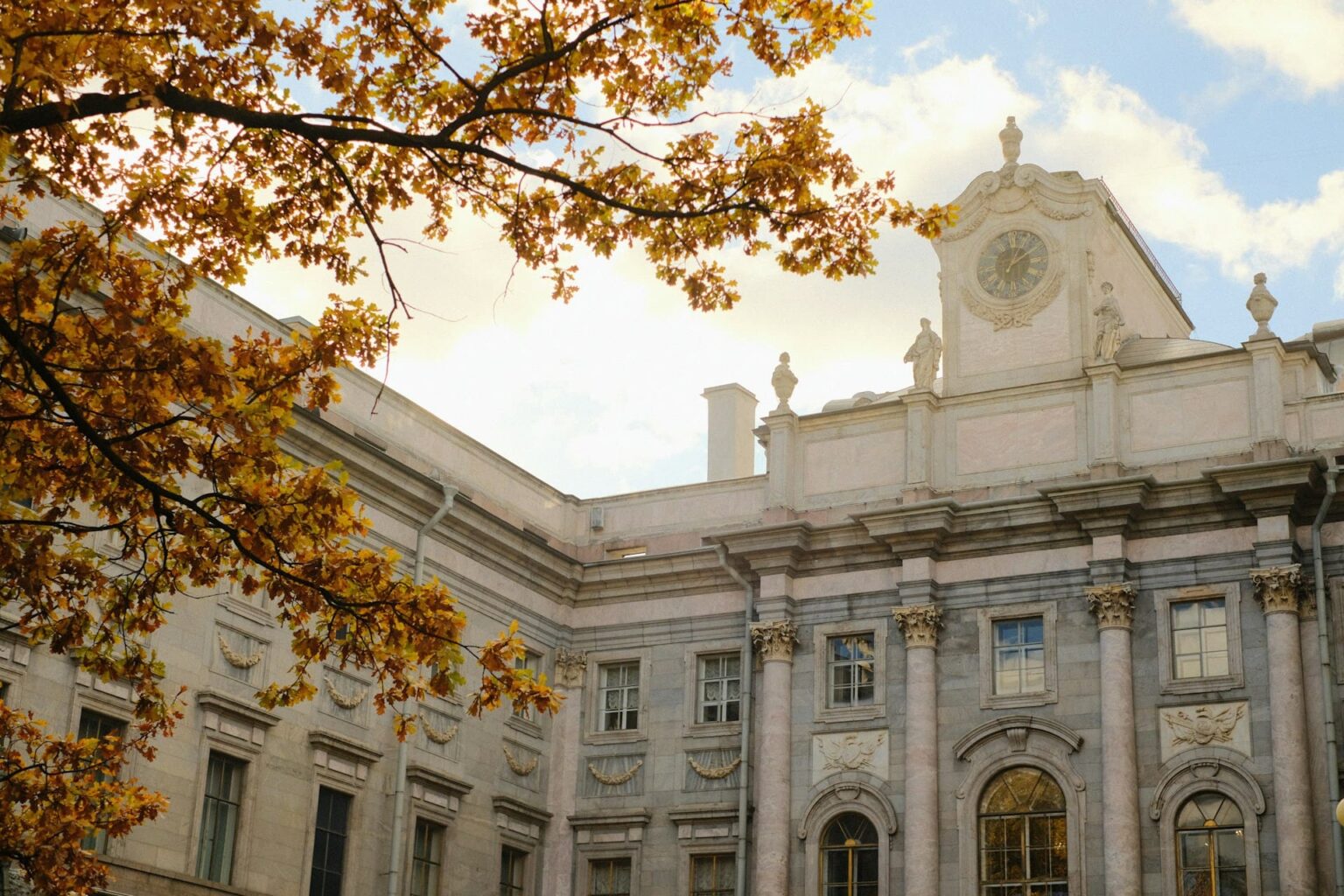 Fachada de um palácio neoclássico com um relógio no alto, atrás de galhos com folhas amarelas de outono