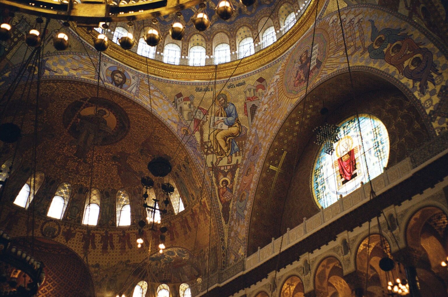 Interior de uma catedral ortodoxa com a cúpula e as paredes cobertas de mosaicos dourados, lustres e um vitral redondo