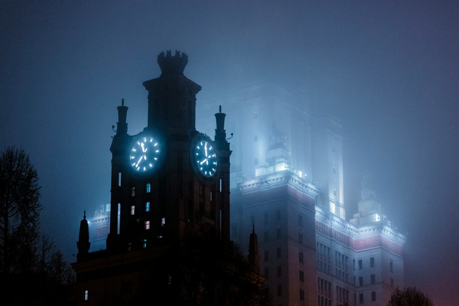 Torre com relógios iluminados diante de um grande edifício, à noite, sob a neblina
