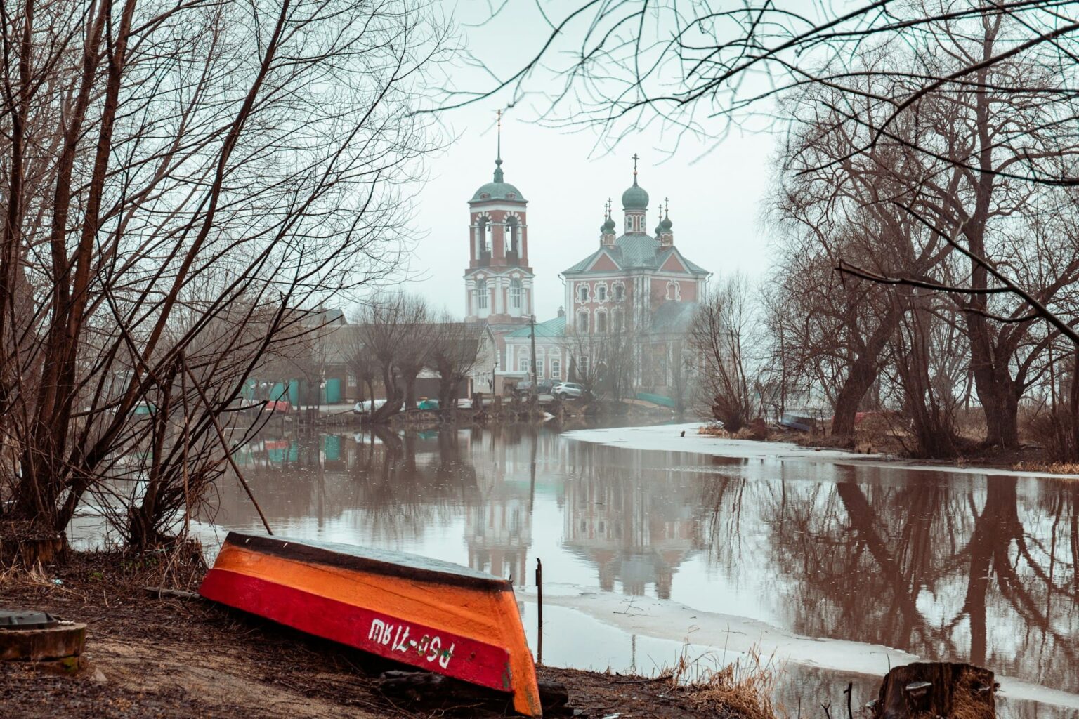 Igreja ortodoxa com campanário à beira de um rio cheio, com um barco virado na margem e árvores sem folhas
