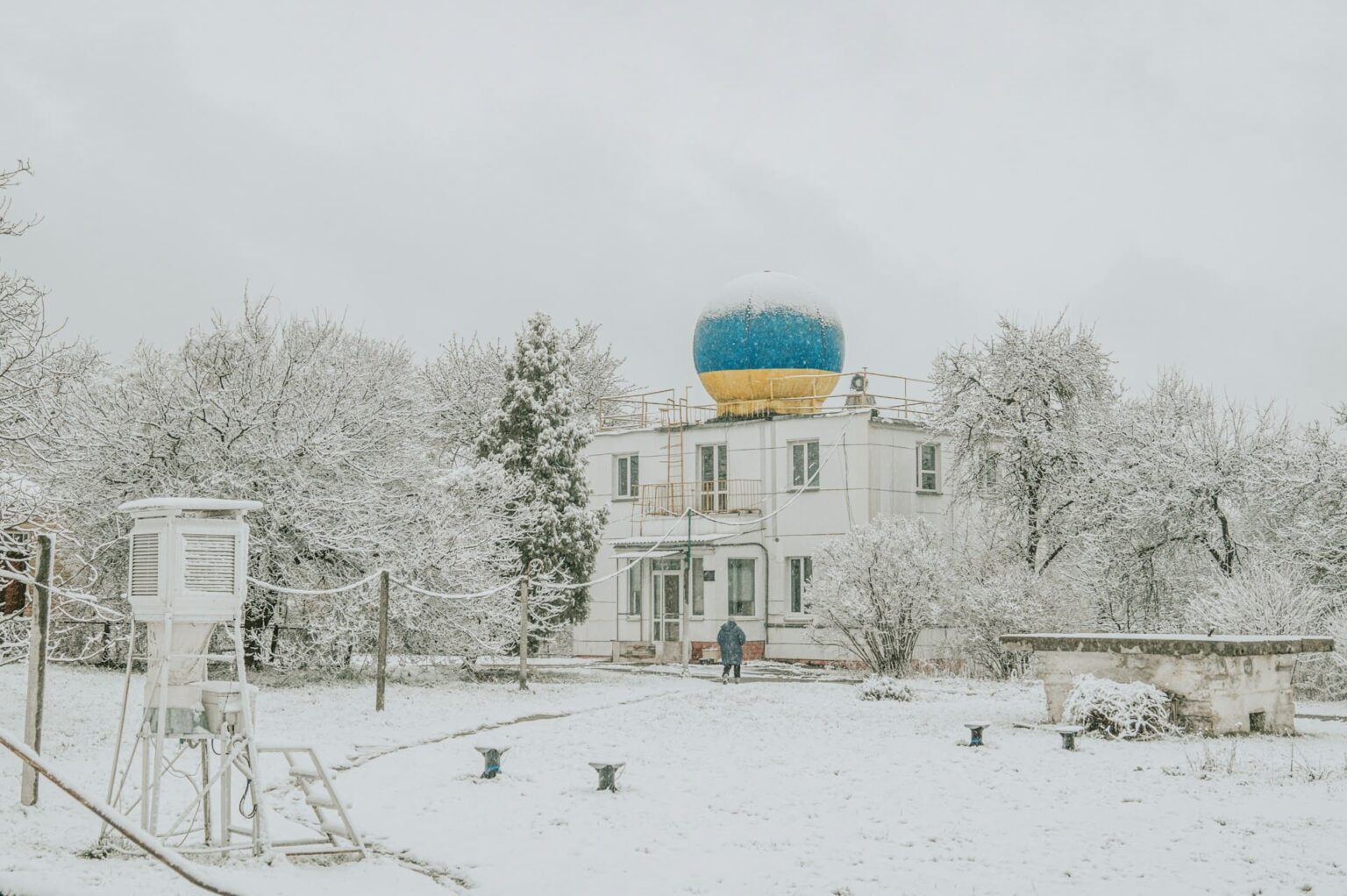 Prédio branco com cúpula azul e amarela, cercado de árvores e neve, com um abrigo meteorológico em primeiro plano