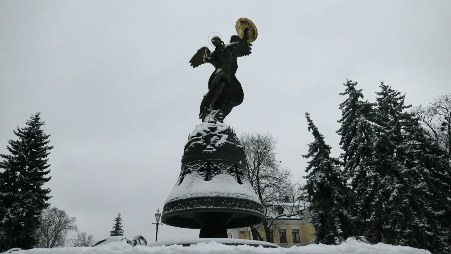 Estátua de um anjo com escudo dourado sobre um grande sino coberto de neve, entre pinheiros