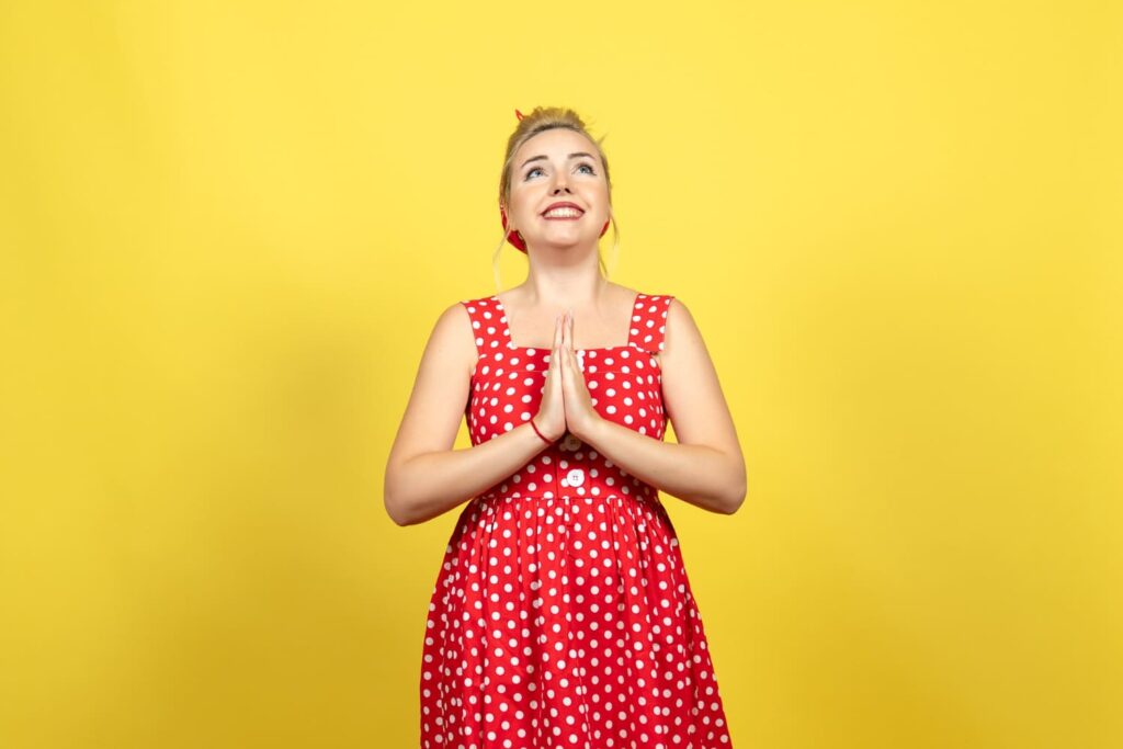 Mulher de vestido vermelho de bolinhas, com as mãos juntas em gesto de súplica, sobre fundo amarelo