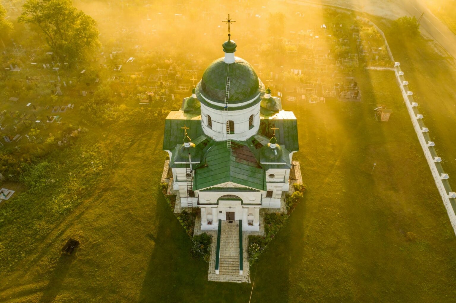 Vista aérea de uma igreja branca com cúpulas verdes, cercada por um campo sob a luz dourada da manhã