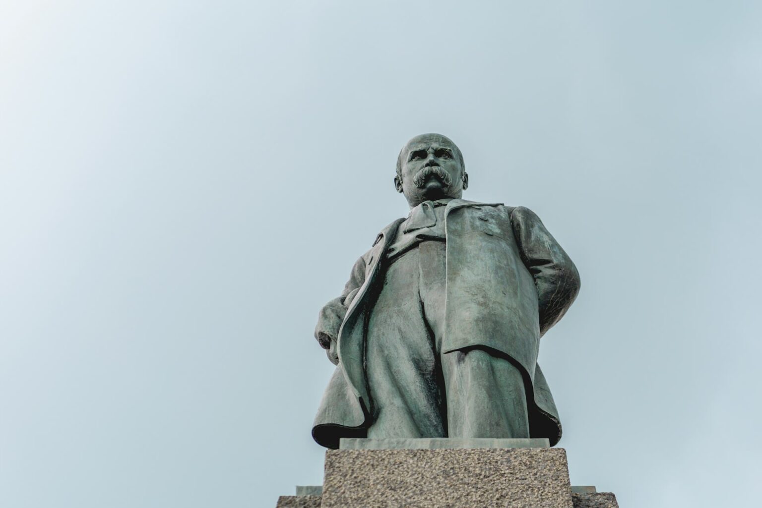 Estátua de bronze de um homem de bigode e casaco longo, sobre um pedestal de granito, vista de baixo